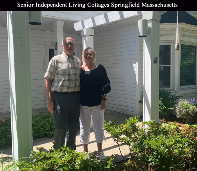 An elderly man and woman standing together outside near a white building with greenery and plants around them. The man is wearing a plaid shirt and dark pants, and the woman is wearing a black top and white pants. They are standing under a white pergola structure.