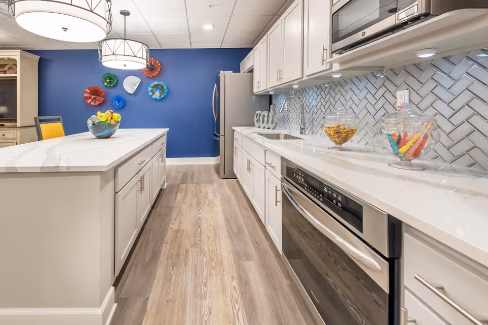 Modern kitchen with white cabinets, a large island with a bowl of fruit, stainless steel appliances including a refrigerator and dishwasher, a white countertop, and a blue accent wall decorated with colorful circular wall art. The floor is light wood, and there are two large pendant lights hanging above the island.