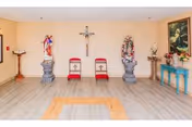 A small chapel or prayer room with two red cushioned chairs placed against a beige wall. Above the chairs is a crucifix. On either side of the chairs are religious statues on pedestals, one of which is adorned with a floral wreath. To the right is a blue table with flowers and a religious statue, and to the left is a wooden lectern. The floor is light wood with a square pattern in the center.