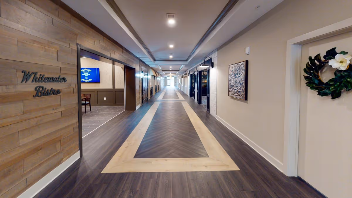A long, well-lit hallway with dark wood flooring featuring a lighter wood inlay pattern. On the left side, there is an entrance to a room labeled 'Whitewater Bistro' with a glimpse of tables and chairs inside. The right side has a door decorated with a green wreath and a framed artwork on the wall. The ceiling has recessed lighting and a tray ceiling design.