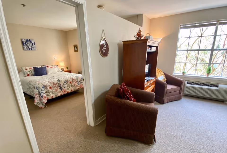 Bright senior living apartment interior with a small living area featuring two armchairs and a TV cabinet by a large window, and a bedroom visible through an open doorway.
