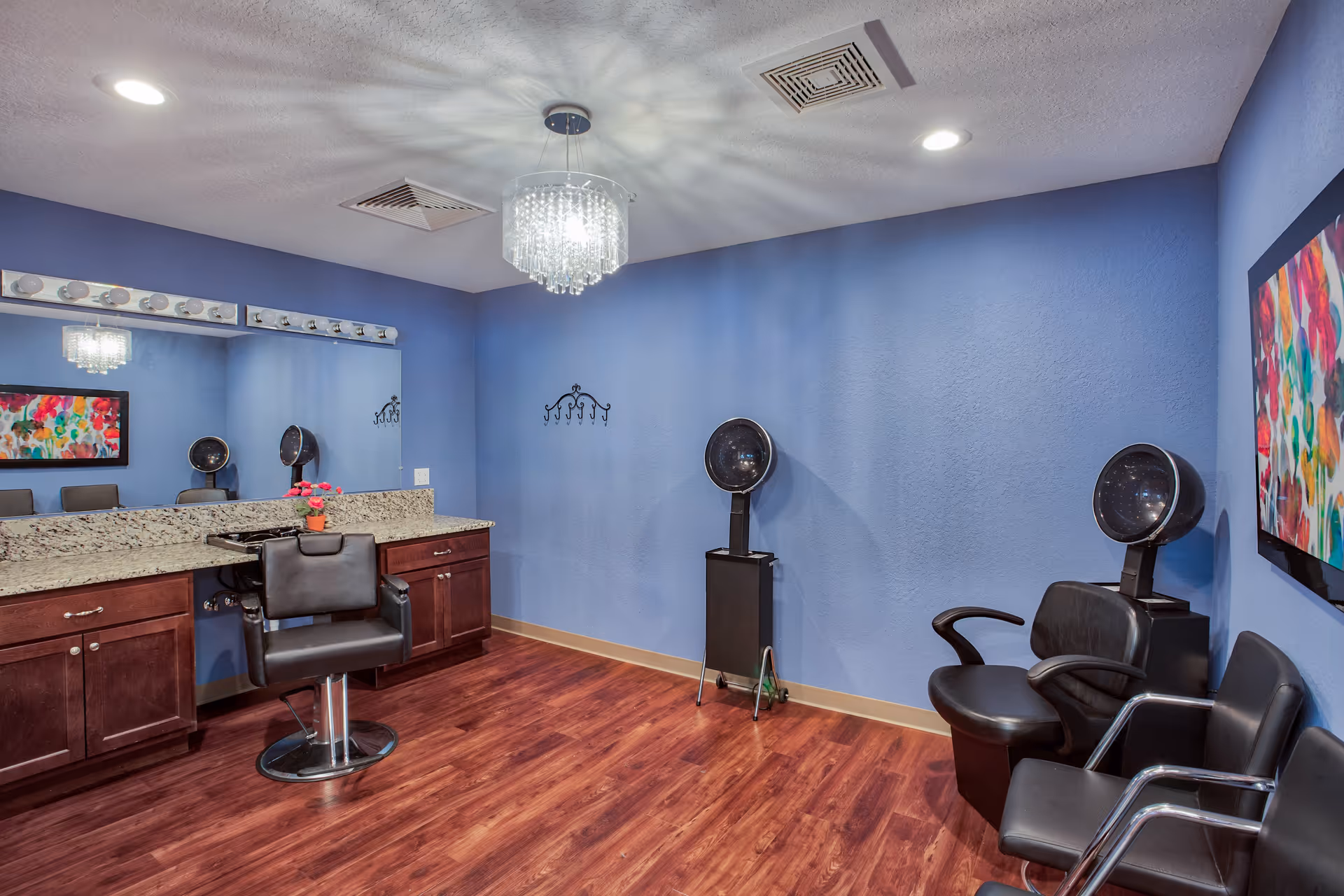 Interior of a salon room with blue walls, wooden floor, a large mirror above a granite countertop with cabinets, a black salon chair, two black hair dryers, and three black waiting chairs. A colorful abstract painting is on the wall.