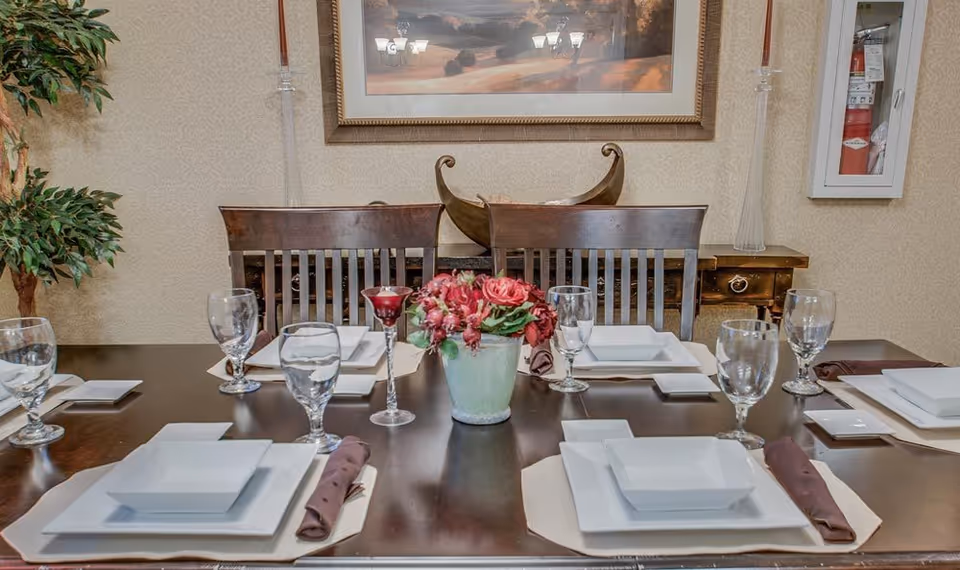 A dining table set for six with white square plates, brown napkins, and clear drinking glasses. A small green vase with red flowers and a red candle holder are placed in the center of the table. Behind the table are wooden chairs, a sideboard with decorative items, a framed landscape painting on the wall, and a fire extinguisher cabinet mounted on the wall to the right.