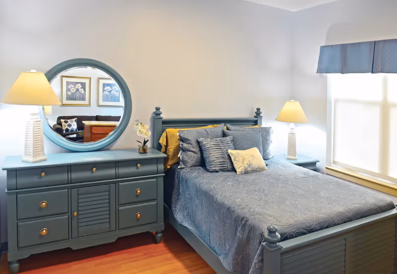 Bedroom with a blue wooden bed and matching dresser topped by a round mirror and table lamps beside a window.