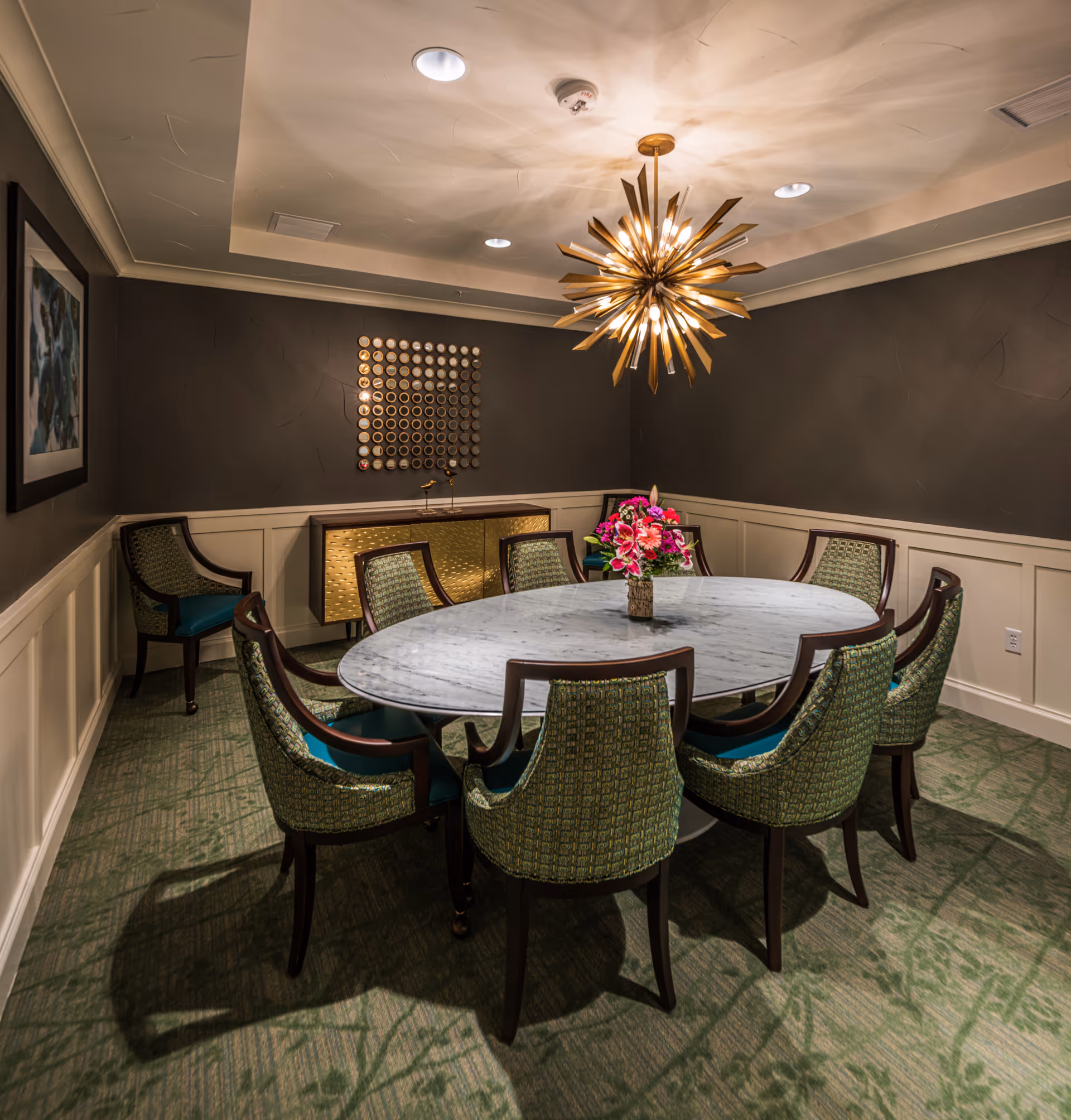 A well-lit dining room with an oval marble table surrounded by eight green upholstered chairs. A modern, starburst chandelier hangs above the table, casting intricate shadows on the ceiling. The walls are painted dark with white wainscoting, and there is a decorative gold sideboard against the back wall with a circular wall art piece above it. A vase with bright pink flowers is placed in the center of the table.