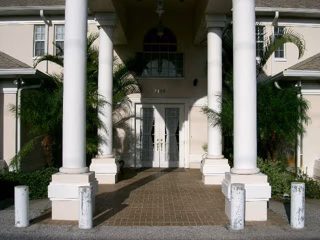Entrance of a building with large white columns, double glass doors, and surrounding greenery including palm trees.