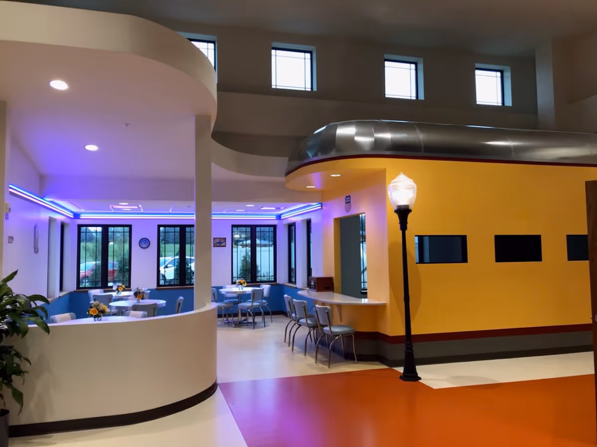Interior of a senior living facility featuring a dining area with round tables and chairs, decorated with small flower arrangements. The space has large windows, a curved white partition, and a yellow wall with a counter and chairs. A streetlamp-style light fixture is also visible inside the room.
