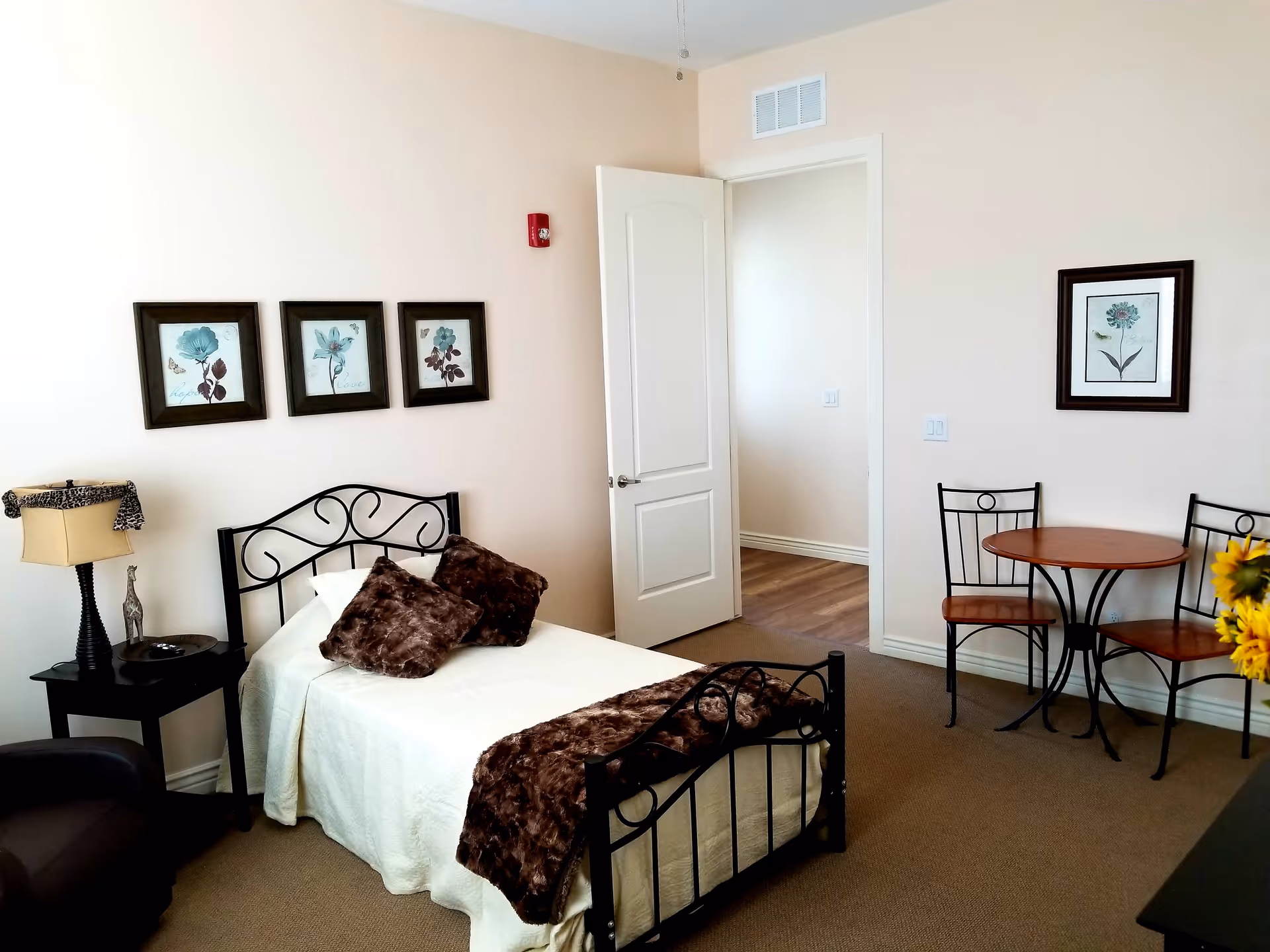 A cozy bedroom with a single bed featuring a black metal frame, white bedding, and brown fur pillows and throw. There is a black side table with a lamp and decorative figurine next to the bed. On the wall above the bed are three framed floral artworks. Across from the bed is a small round wooden table with two matching chairs, and a framed flower picture on the wall. The room has beige walls, a carpeted floor, and an open white door leading to another room with wooden flooring.