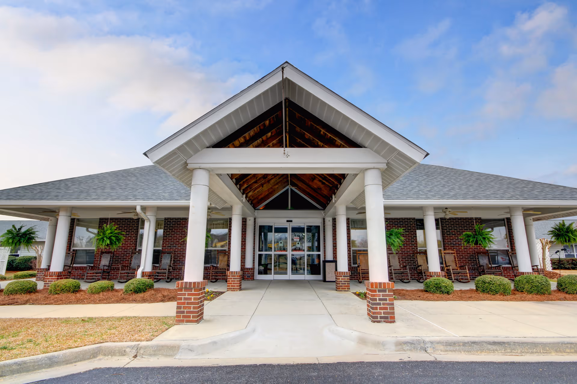 Front exterior view of a building with a covered entrance supported by white columns with brick bases. The building has a gray shingled roof and brick walls. There are hanging green plants and rocking chairs on the porch area under the roof. The sky is partly cloudy.