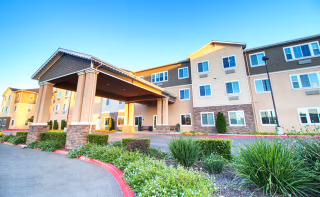 Exterior view of a multi-story senior living facility building with a covered entrance, landscaped greenery, and clear blue sky.