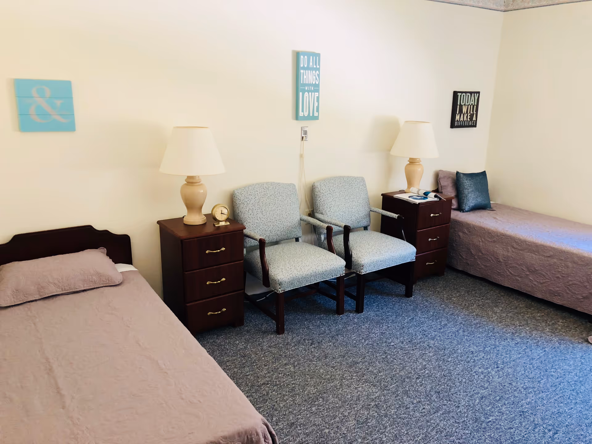 A simple, clean bedroom with two single beds covered in light purple bedspreads. Between the beds are two wooden nightstands, each with a beige lamp. Two upholstered chairs with wooden arms are placed side by side between the nightstands. The walls are light-colored and decorated with three small framed inspirational quotes.