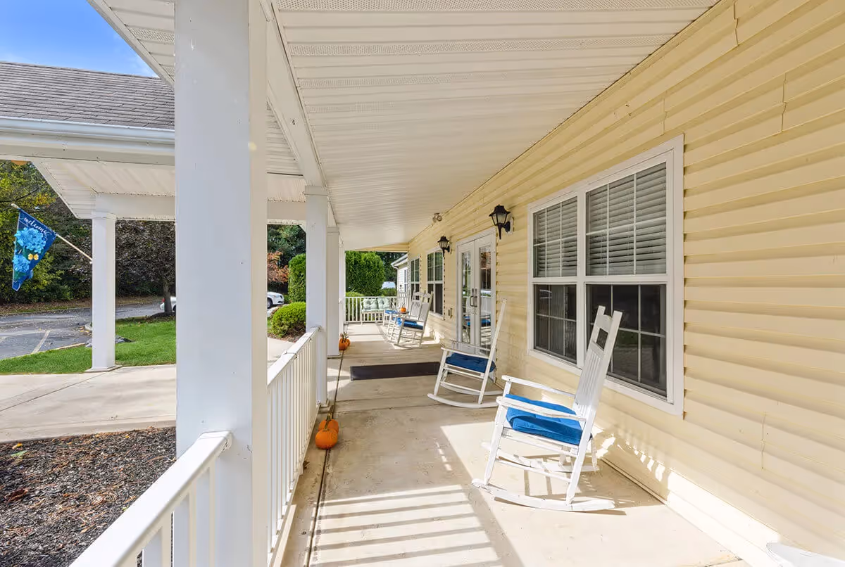 Covered porch area of a facility with white rocking chairs that have blue cushions, beige siding walls, white railing, and several pumpkins placed along the walkway. There are windows with white blinds and black wall-mounted lantern lights. A blue flag with a floral design is visible near the parking area.