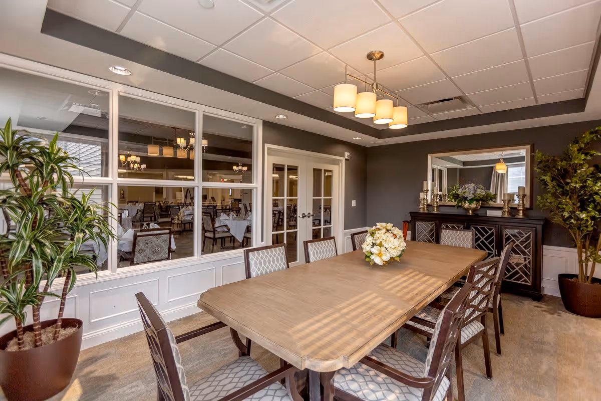 A well-lit dining room with a long wooden table surrounded by eight chairs with patterned cushions. A floral centerpiece is placed on the table. The room has dark gray walls, a large mirror above a sideboard with decorative items, and two potted plants in the corners. Through glass windows and double doors, another dining area with tables and chairs is visible.