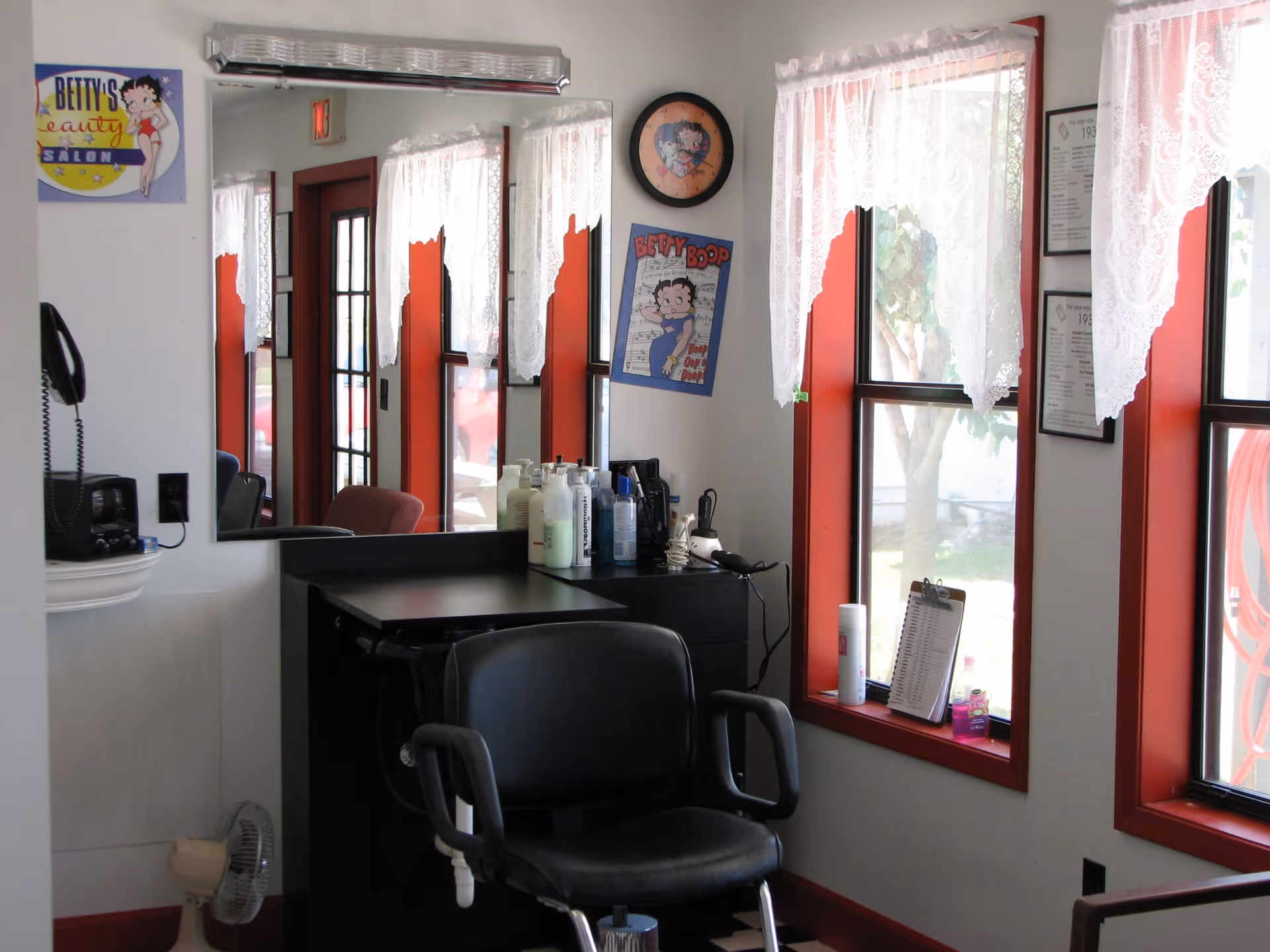 Interior of a small beauty salon with a black salon chair in front of a counter holding various hair care products. The room has large windows with white lace curtains and red trim. A mirror on the wall reflects part of the room, and there are Betty Boop themed decorations on the walls.