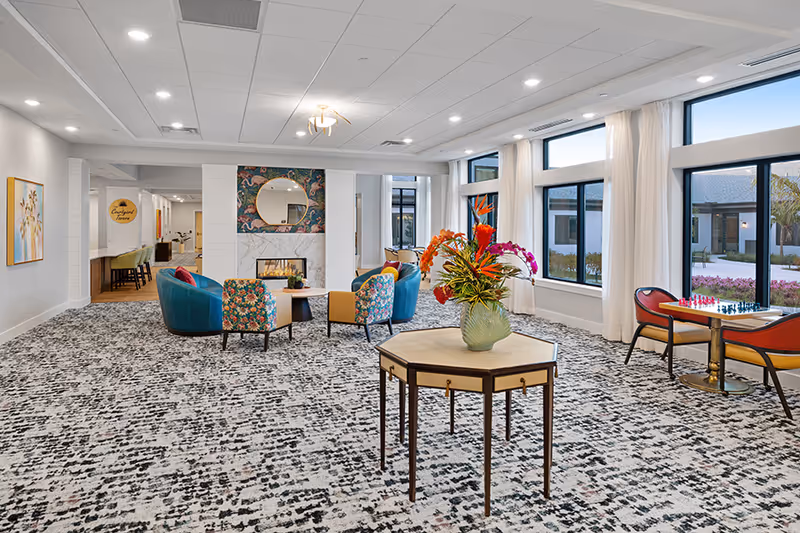 A bright and spacious common area with large windows letting in natural light. The room features a patterned carpet, a central table with a vase of colorful flowers, and several seating arrangements including floral and solid-colored chairs around a fireplace with a round mirror above it. There is also a small table with two chairs near the windows, set up for a chess game. The walls are light-colored with some artwork, and the ceiling has recessed lighting.