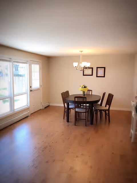 A bright dining room with light wooden flooring, a round dark wooden table surrounded by six matching chairs, a vase with yellow flowers on the table, two framed pictures on the beige wall, a chandelier with five lights hanging above the table, and large windows with blinds letting in natural light next to a door.