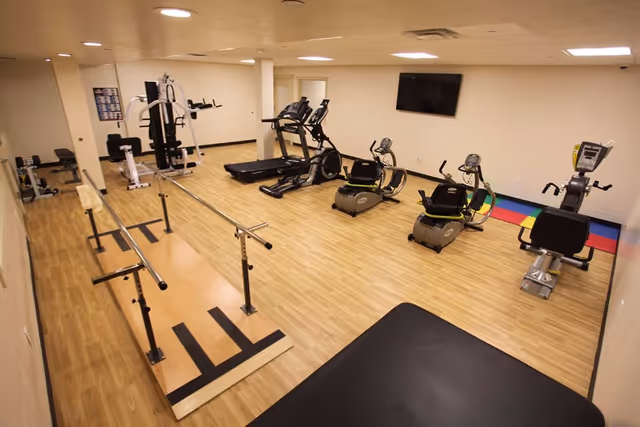 A fitness room with wooden flooring featuring exercise equipment including two treadmills, two recumbent bikes, a rowing machine, a multi-station gym machine, and a ballet barre. A flat-screen TV is mounted on the wall, and there is a colorful mat in the corner.