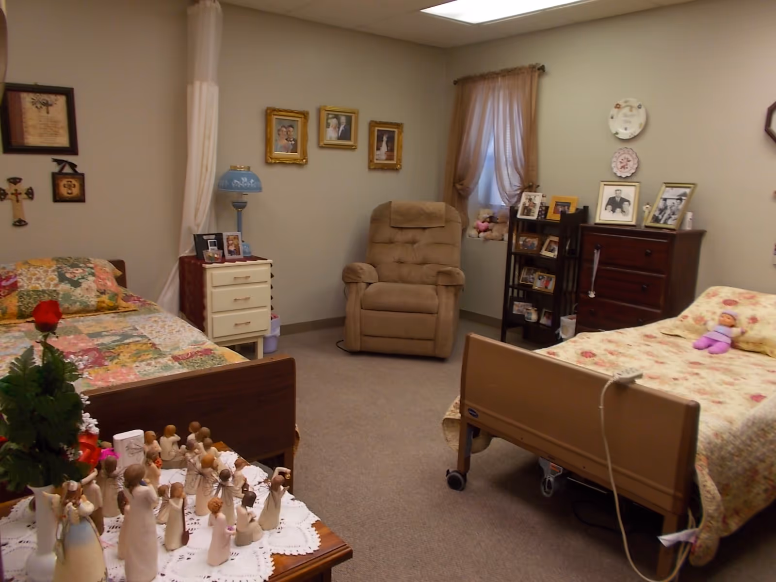 Shared assisted-living bedroom with two beds, a recliner, dressers, and personal photos and decorations.