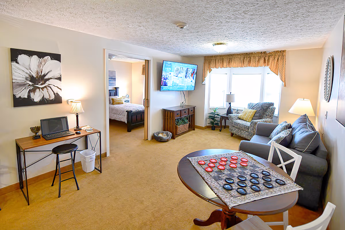 Bright furnished living room with a round table and checkers set, sofa and armchair by a window, a TV on the wall, and a small desk with a laptop opening into a bedroom.