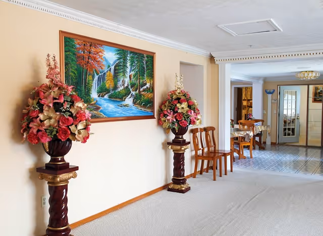 Interior view of a senior living facility hallway with beige walls and carpeted floor. Two ornate flower arrangements in vases on decorative pedestals flank a colorful landscape painting of a waterfall and forest. Wooden chairs are lined up along the wall, and a dining area with tables and chairs is visible in the background near a tiled floor and glass door.