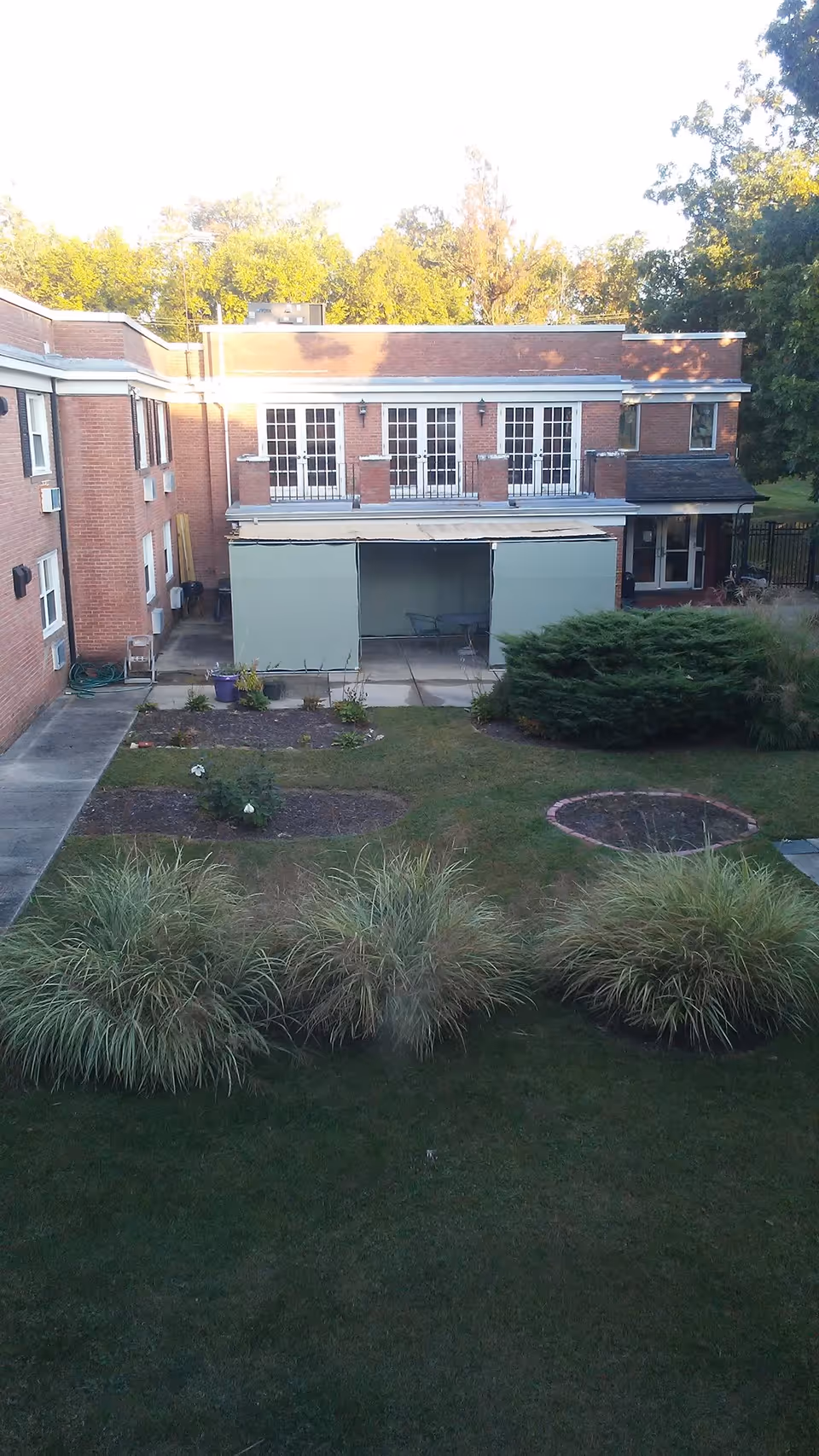 View of the outdoor garden area at King David Nursing and Rehabilitation Center, showing a grassy lawn with ornamental grasses and small flower beds in front of a two-story brick building with multiple windows and a covered patio area.