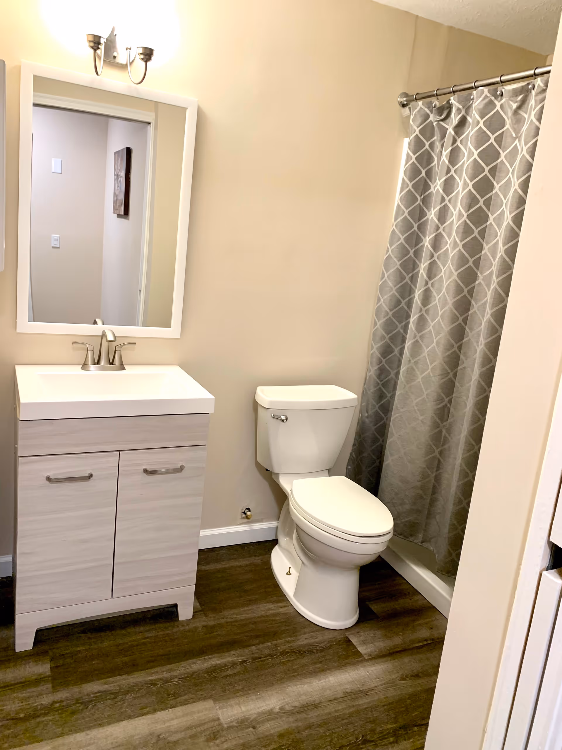 A clean bathroom with a white toilet, a light wood vanity with a white countertop and a silver faucet, a rectangular mirror with a white frame above the vanity, a silver light fixture with two bulbs above the mirror, and a shower with a gray and white patterned curtain.