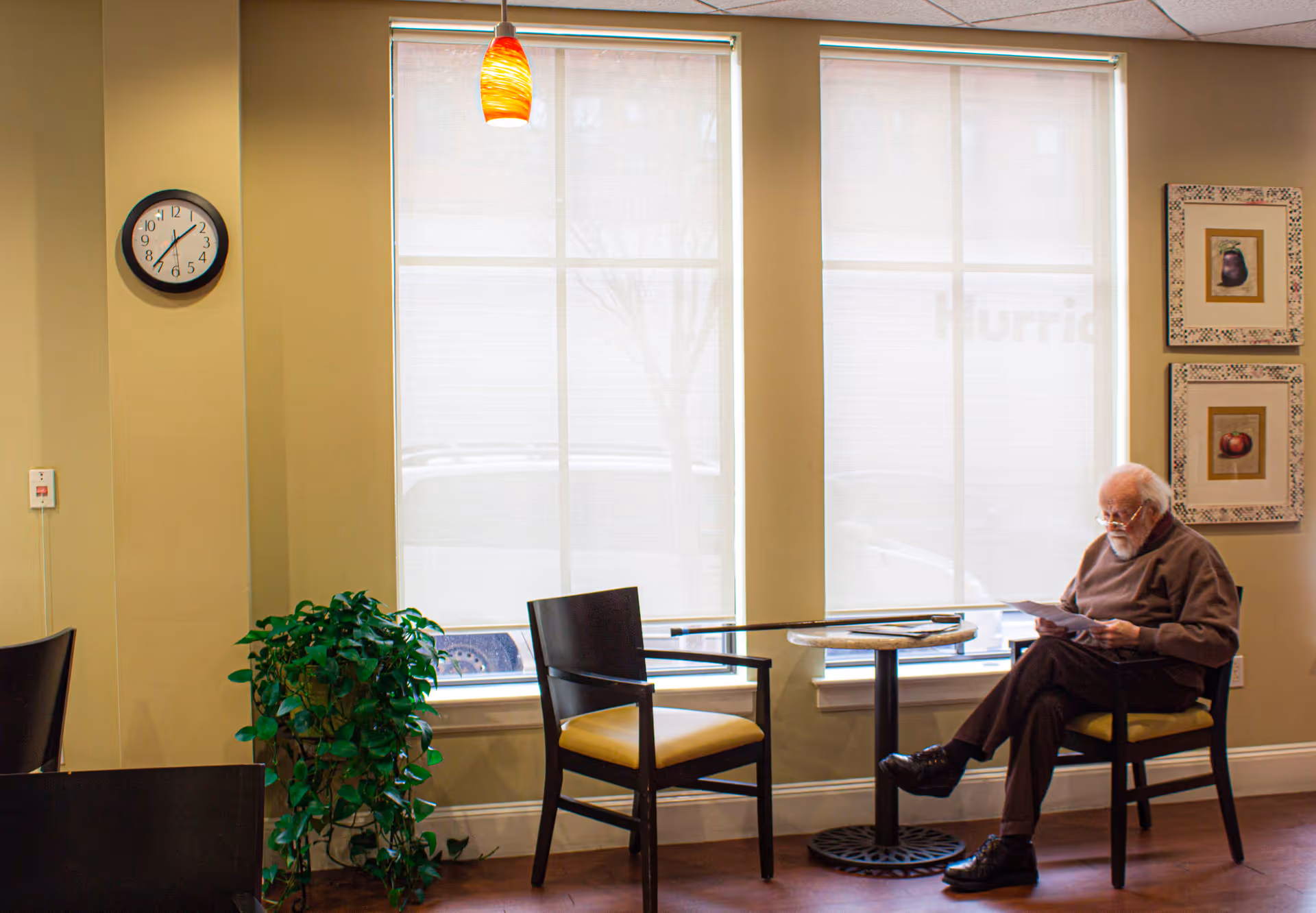 An elderly man with white hair and beard is sitting on a chair at a small round table in a well-lit room with large windows covered by blinds. The man is reading a document. The room has beige walls, a clock on the wall, two framed pictures, a green potted plant, and a hanging orange pendant light.