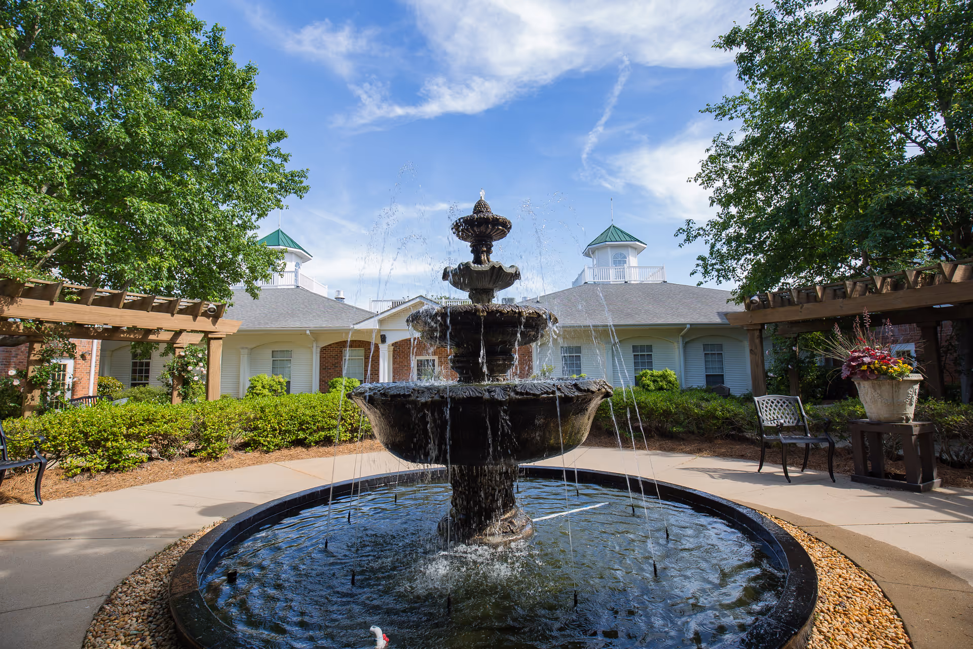 A multi-tiered water fountain in the center of a circular pond surrounded by a paved walkway, with green bushes, trees, and a building with white siding and green-roofed cupolas in the background under a partly cloudy blue sky.