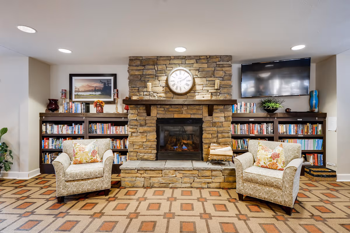 Cozy living room area with a stone fireplace in the center, flanked by two bookshelves filled with books. Above the fireplace is a round clock and two candle holders. Two patterned armchairs with floral cushions are placed on either side of the fireplace. A flat-screen TV is mounted on the wall to the right. The room has a patterned carpet and recessed ceiling lights.