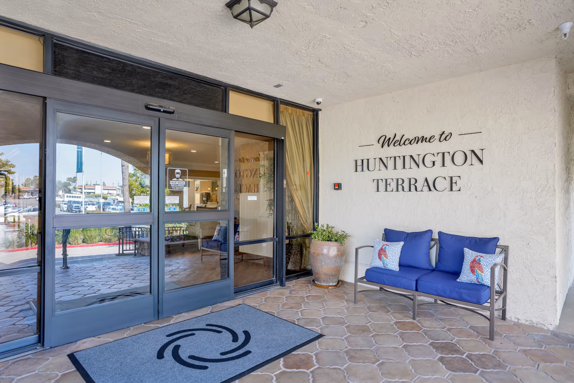 Entrance to Huntington Terrace with automatic glass doors, a blue-cushioned bench, potted plant, and a 'Welcome to Huntington Terrace' wall sign.