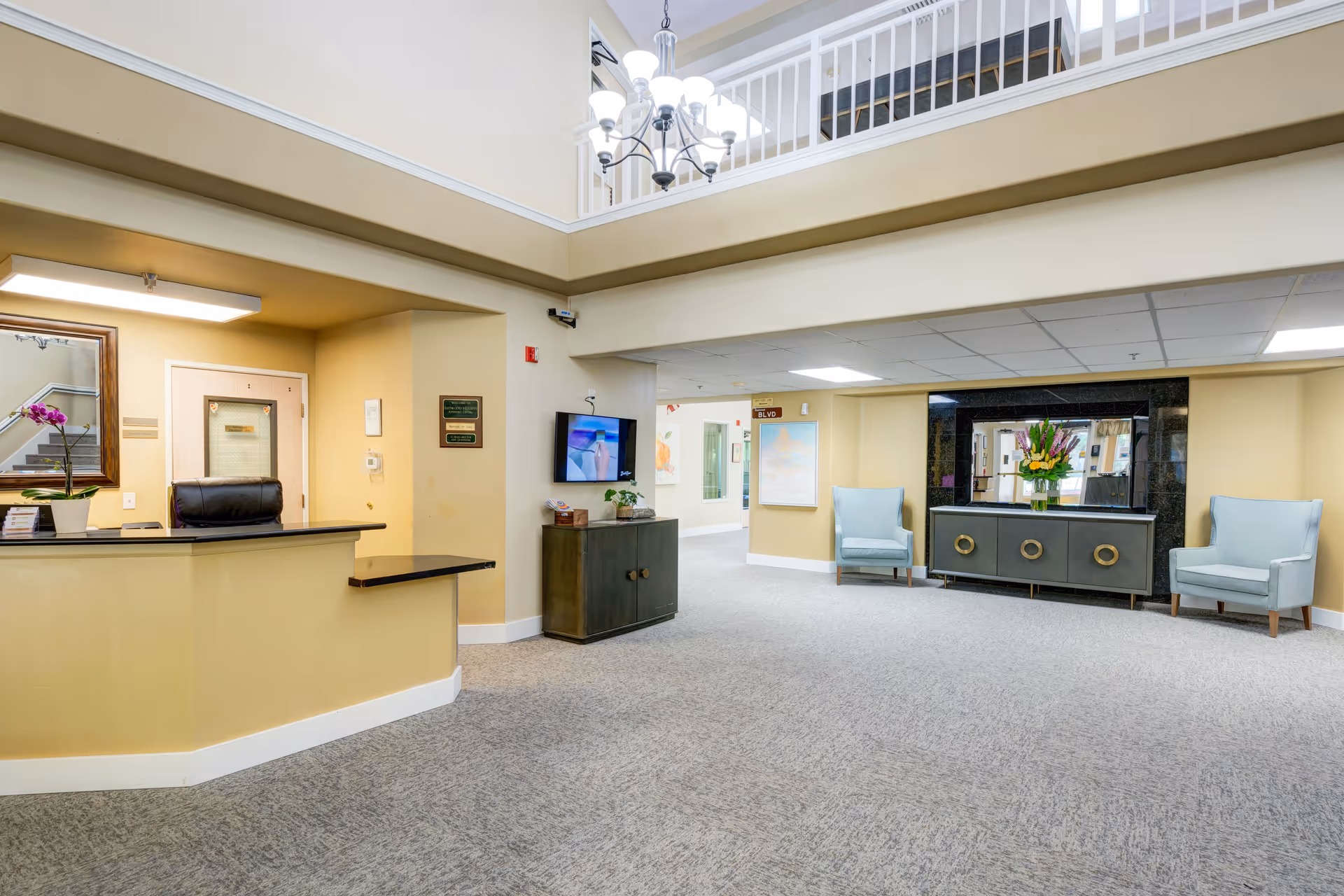Interior view of a senior living facility lobby area with a reception desk on the left, a TV mounted on the wall, two light blue armchairs, a large mirror above a cabinet with a flower vase, and a chandelier hanging from the ceiling with a second-floor railing visible above.