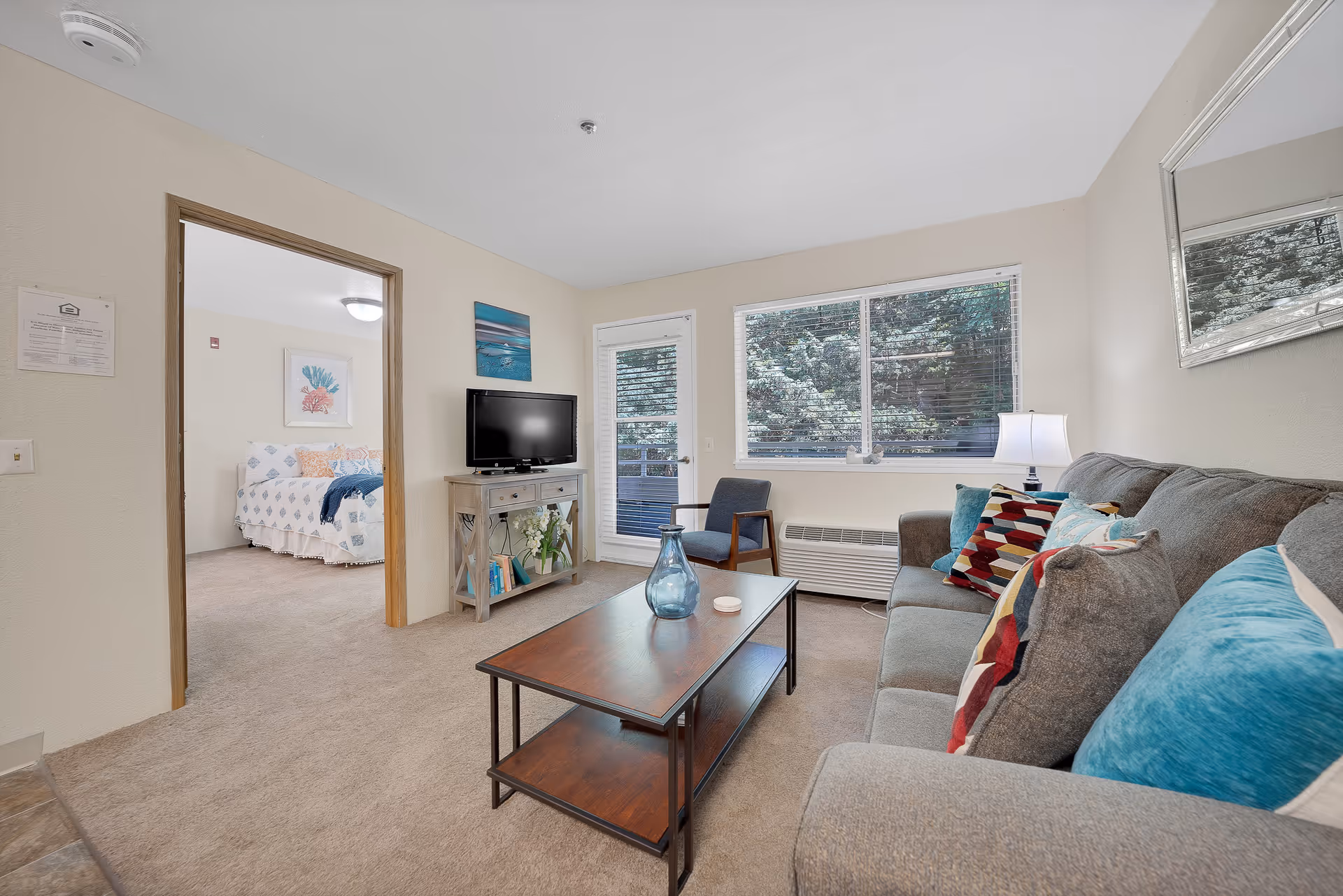 A cozy living room in a senior living facility with a gray sofa adorned with colorful pillows, a wooden coffee table with a blue vase, a single armchair, a TV on a wooden stand, and a large window with blinds showing greenery outside. A doorway leads to a bedroom with a neatly made bed and framed artwork on the wall.