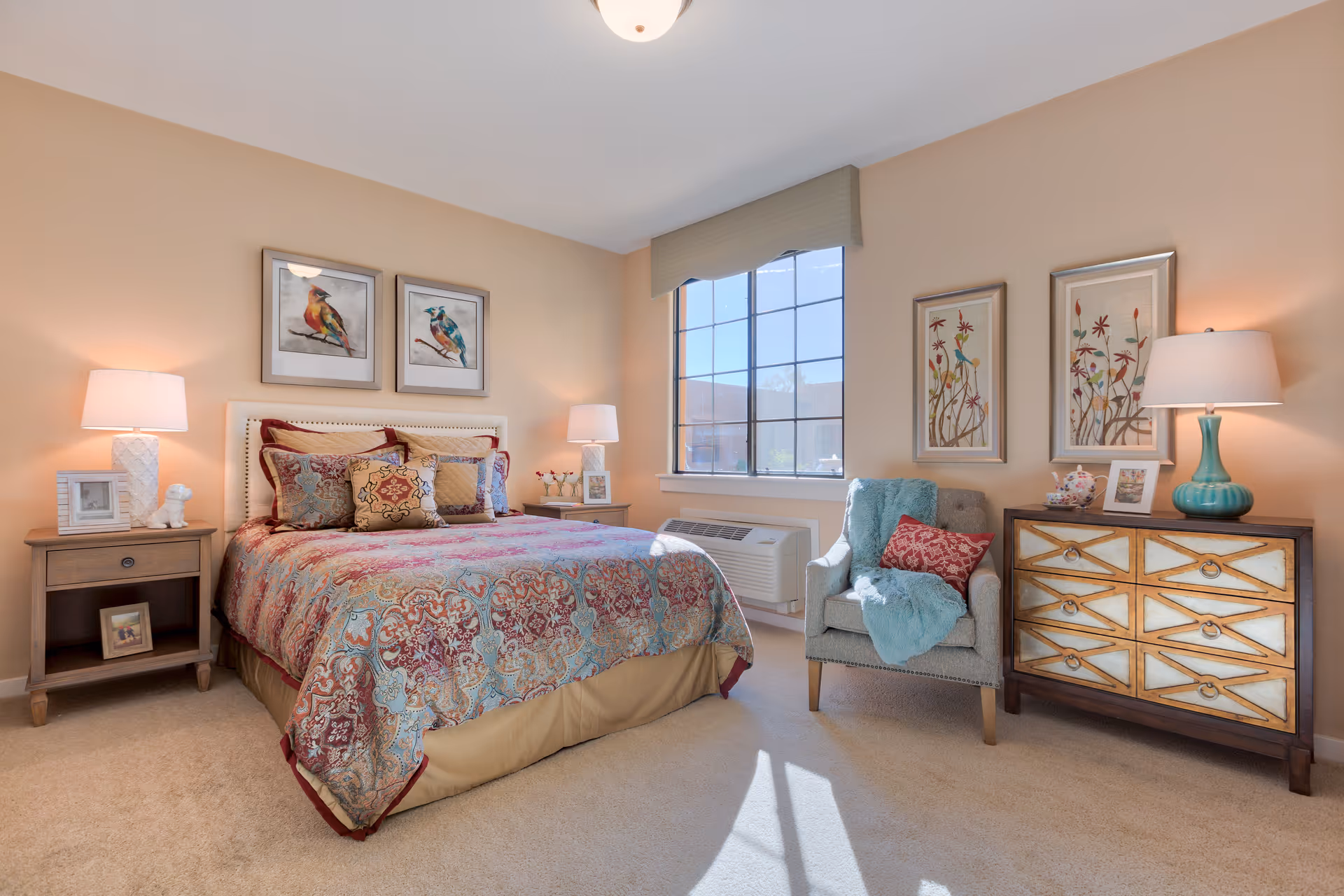 Sunny, well-decorated bedroom with a patterned bed, nightstands and lamps, an armchair, and a dresser beneath a window.