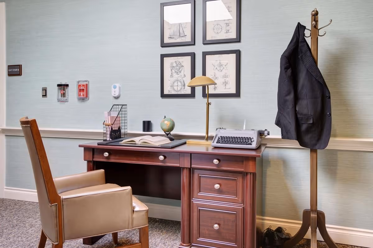 A wooden desk and chair in a small office-like corner with a typewriter, globe, lamp, framed prints on the wall, and a coat on a stand.