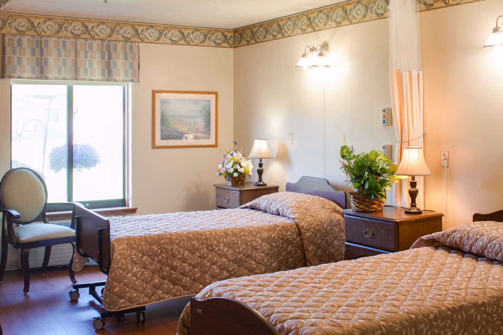 Two twin beds with quilted bedspreads, nightstands with lamps and plants, a chair, and a window in a senior living bedroom.