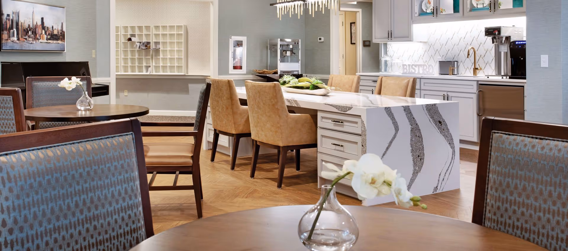 A modern dining area in a senior living facility featuring a large white marble island with beige chairs, round wooden tables with patterned chairs, a small vase with white flowers on the table in the foreground, and a kitchenette with white cabinets and a coffee machine in the background.