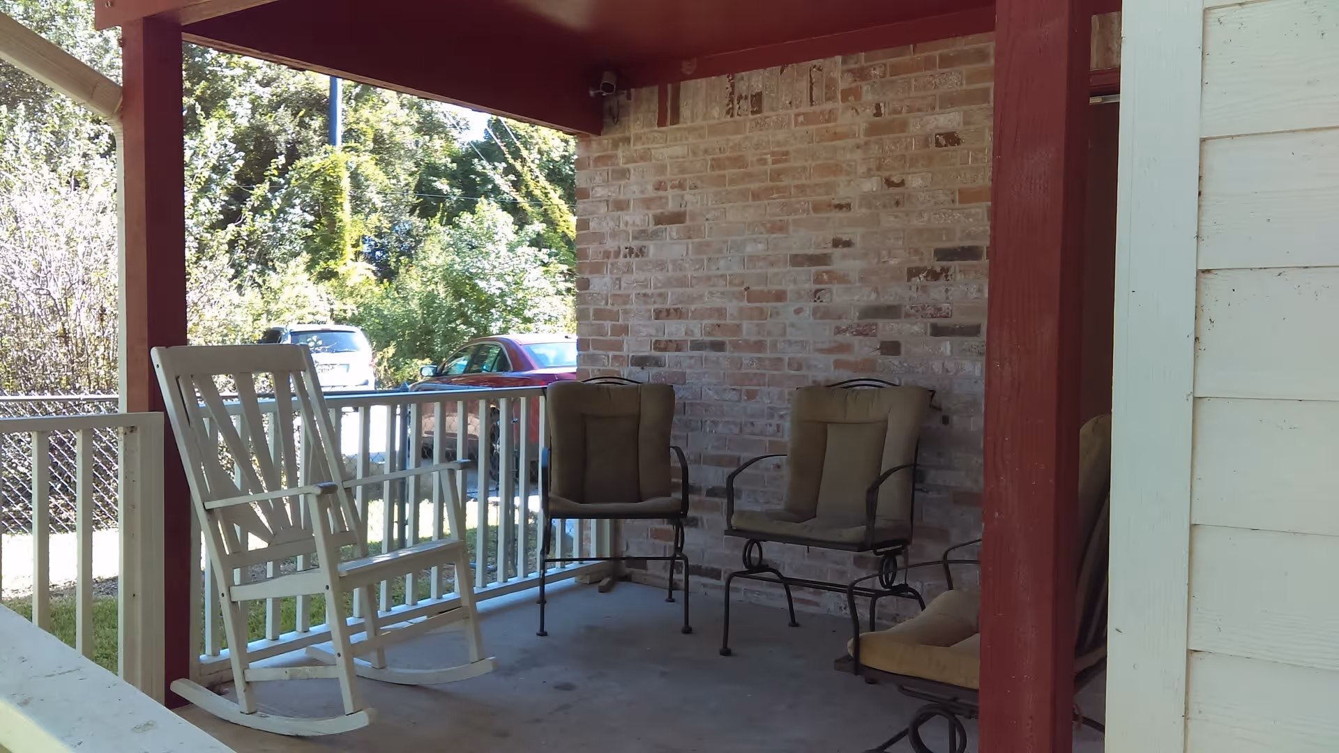 Covered front porch with a white rocking chair and several cushioned metal patio chairs against a brick wall.