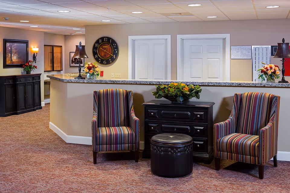 Interior view of a reception area in a senior living facility with a granite countertop desk, two striped armchairs, a black ottoman, a black chest with a flower arrangement, wall clock, lamps, framed artwork, and beige walls and carpet.