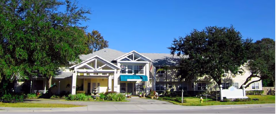 Exterior front view of a two-story senior living facility building with a covered entrance, surrounded by green trees and landscaping under a clear blue sky.