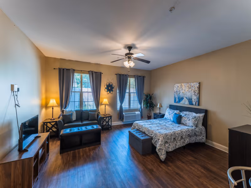 A spacious senior living bedroom with a bed featuring floral bedding, a cushioned bench at the foot of the bed, a small sofa with an ottoman, two windows with gray curtains, two side tables with lamps, a ceiling fan with lights, a TV on a wooden stand, and wood flooring.