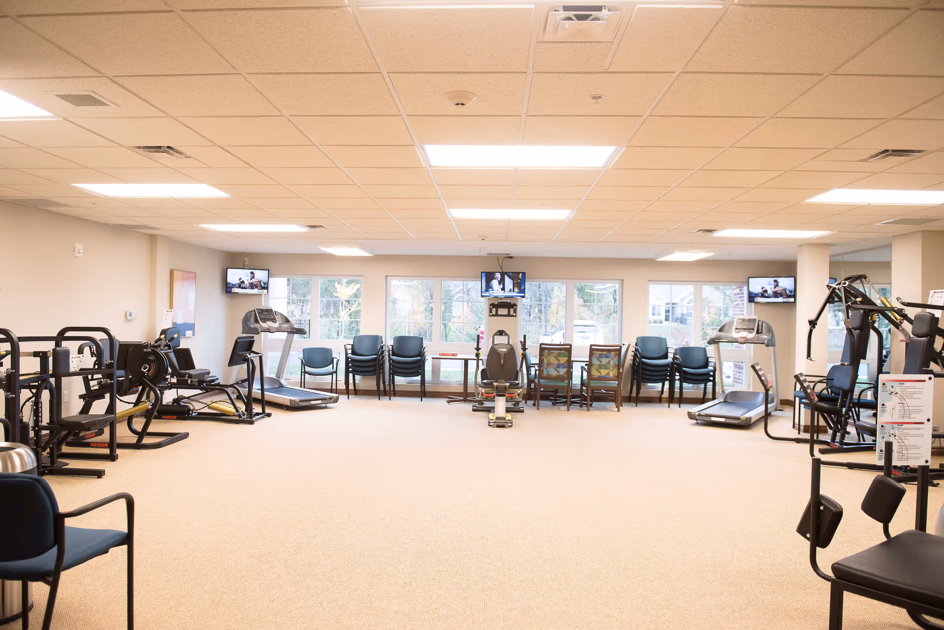 A spacious fitness room in a senior living facility with various exercise equipment including treadmills, weight machines, and stationary bikes. Several chairs are lined up against the windows, and three televisions are mounted on the walls. The room has a beige carpet and a drop ceiling with fluorescent lighting.