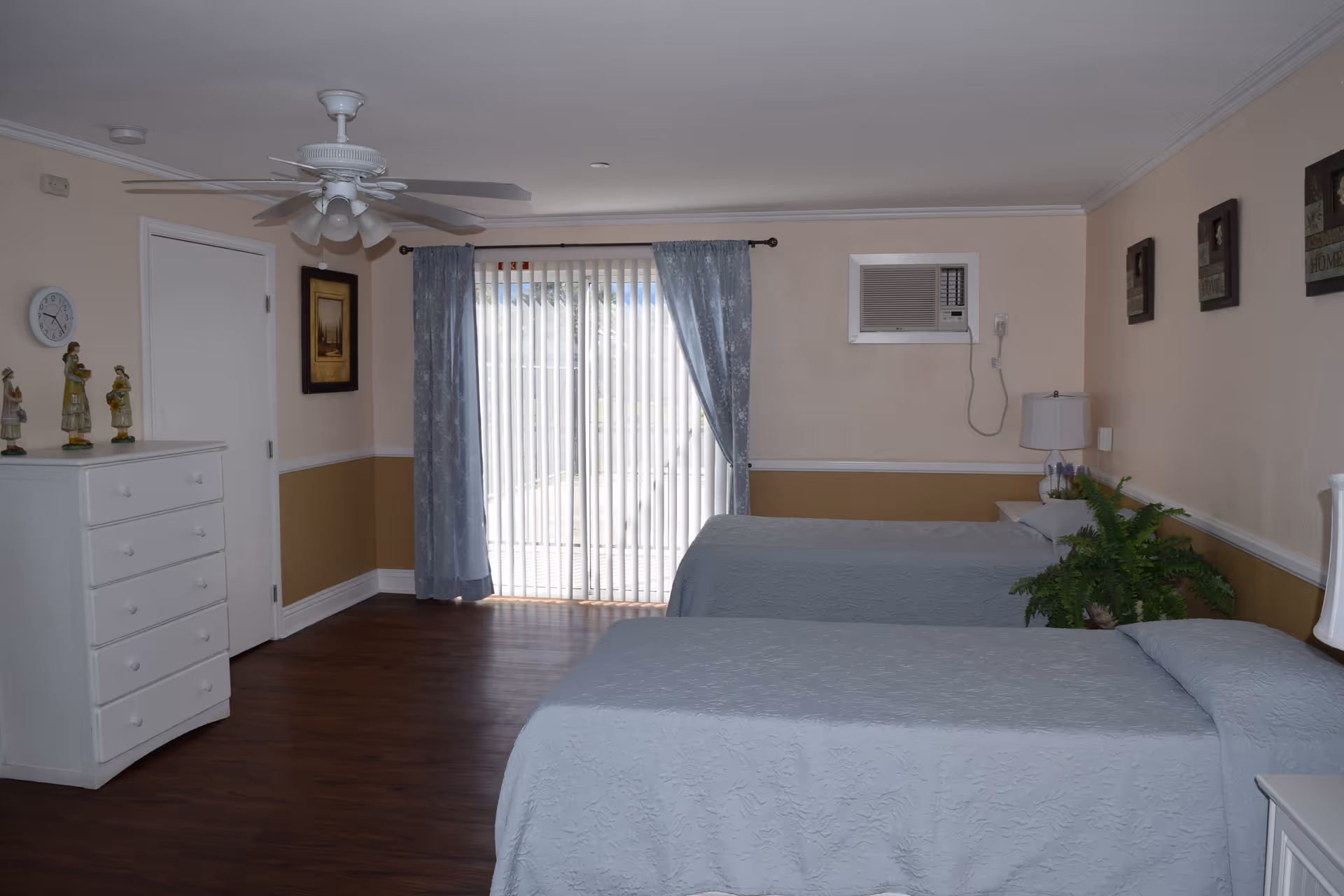 A bedroom with two twin beds covered in light gray bedspreads. The room has wooden flooring, a white ceiling fan, a white dresser with decorative figurines, and a sliding glass door with vertical blinds and blue curtains. There is an air conditioning unit installed on the wall above a nightstand with a lamp and a green plant. Three framed pictures hang on the wall above one of the beds.
