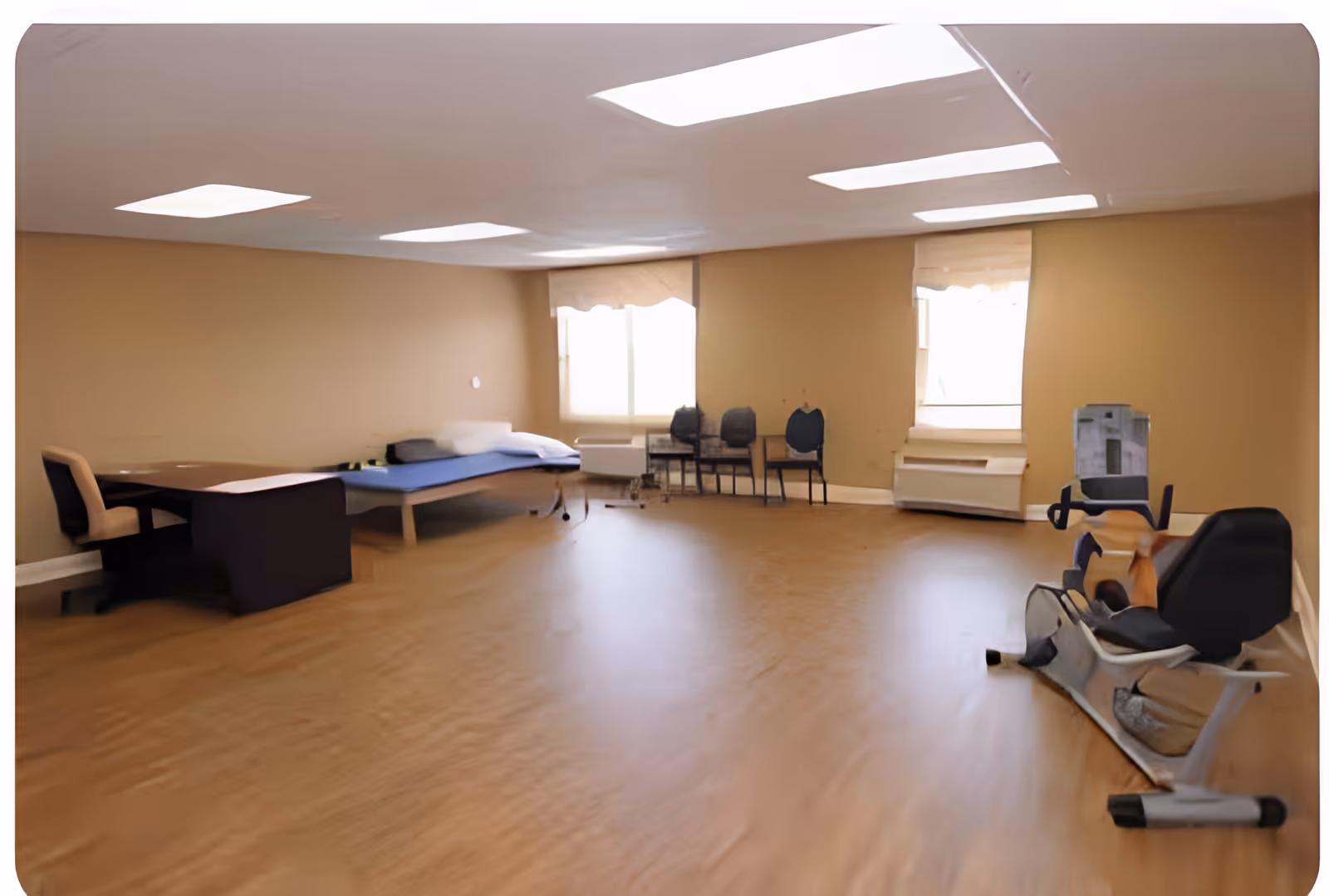 A spacious room with beige walls and wooden flooring featuring a single bed with a blue mattress, a desk with a chair, several stacked chairs near two windows with blinds, and a stationary exercise bike.