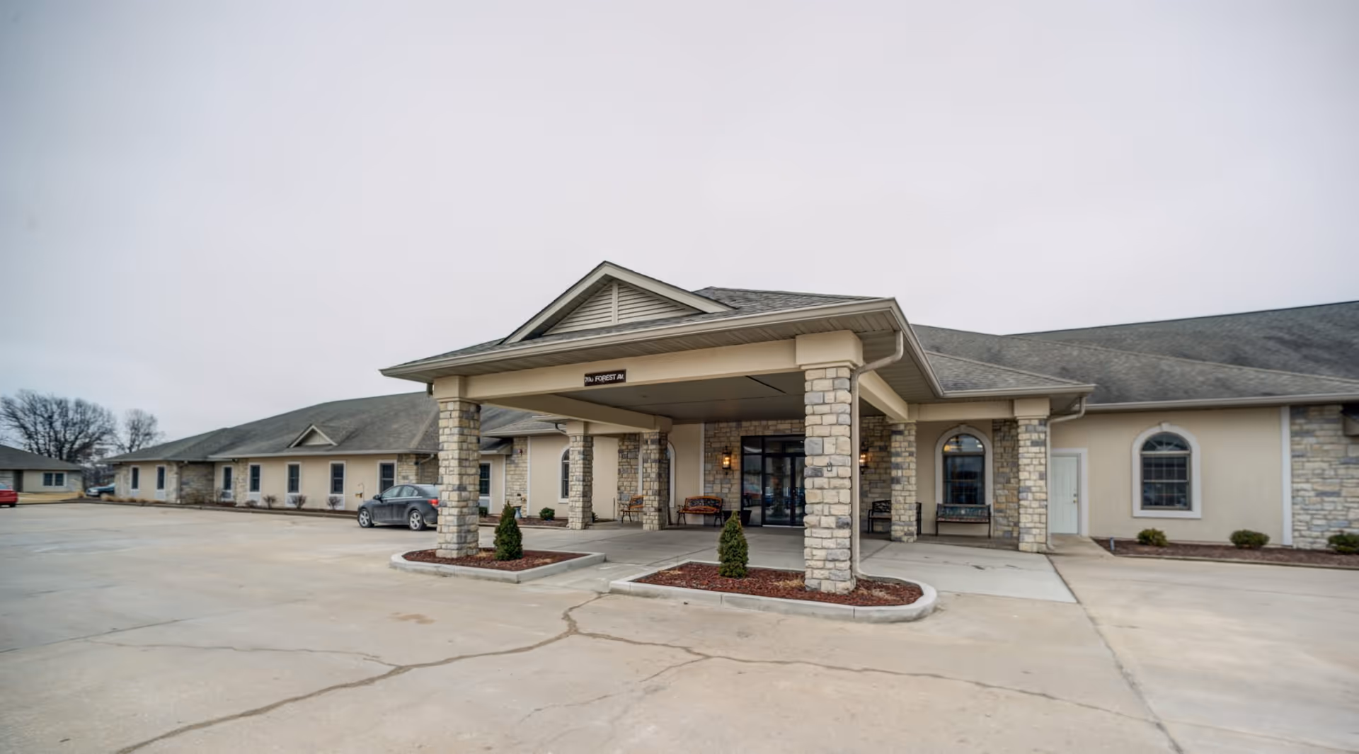 Exterior view of Oak Pointe Assisted Living facility showing a single-story building with stone pillars supporting a covered entrance. There are benches near the entrance and a few cars parked in the parking lot under an overcast sky.