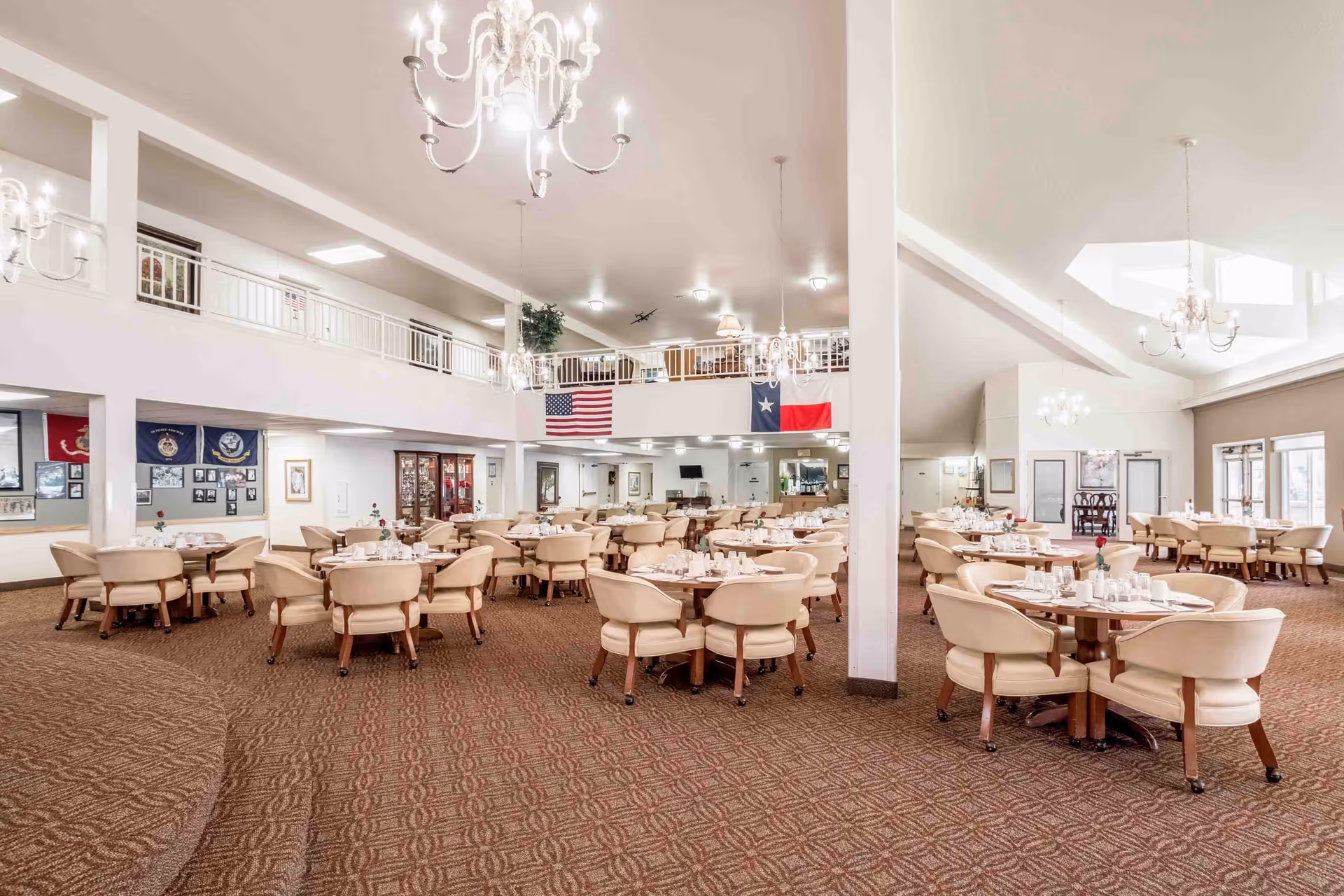 Spacious dining room with multiple round tables surrounded by beige cushioned chairs on wheels. The room has a high ceiling with chandeliers and large windows allowing natural light. American and Texas flags hang on the upper balcony railing. The carpet has a patterned design and the walls are decorated with framed pictures and memorabilia.