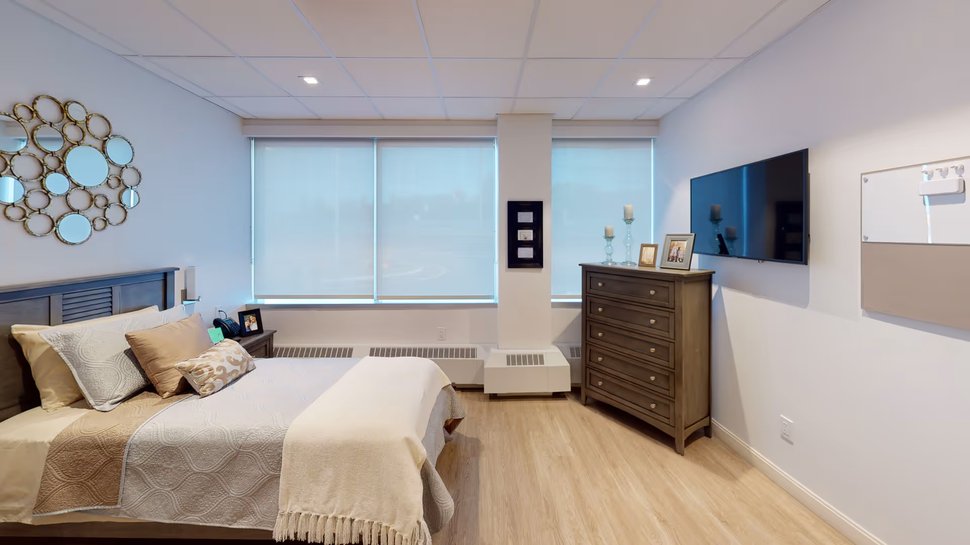 A bright senior living bedroom with a made bed, decorative pillows, a wooden dresser with framed photos and candles, a wall-mounted TV, and large windows with shades.