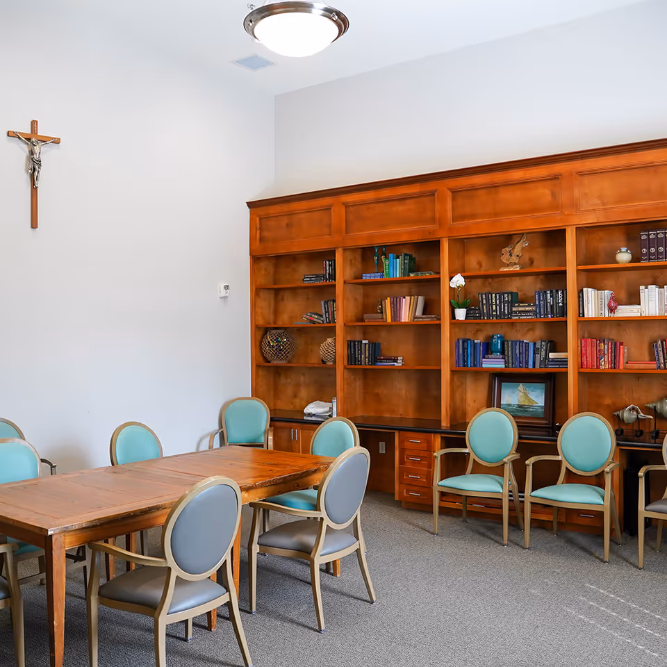 A quiet room with a wooden table surrounded by chairs with light blue and gray cushions. Behind the table is a large wooden bookshelf filled with books, decorative items, and a small framed picture. A crucifix is mounted on the white wall above the table, and a ceiling light fixture is visible.