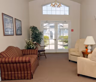 A cozy sitting area in a senior living facility with a striped sofa on the left, two beige armchairs on the right, a wooden side table with a lamp, a potted plant, and a wooden chair near glass double doors leading outside. The room has a high ceiling with a window above the doors and framed pictures on the wall.
