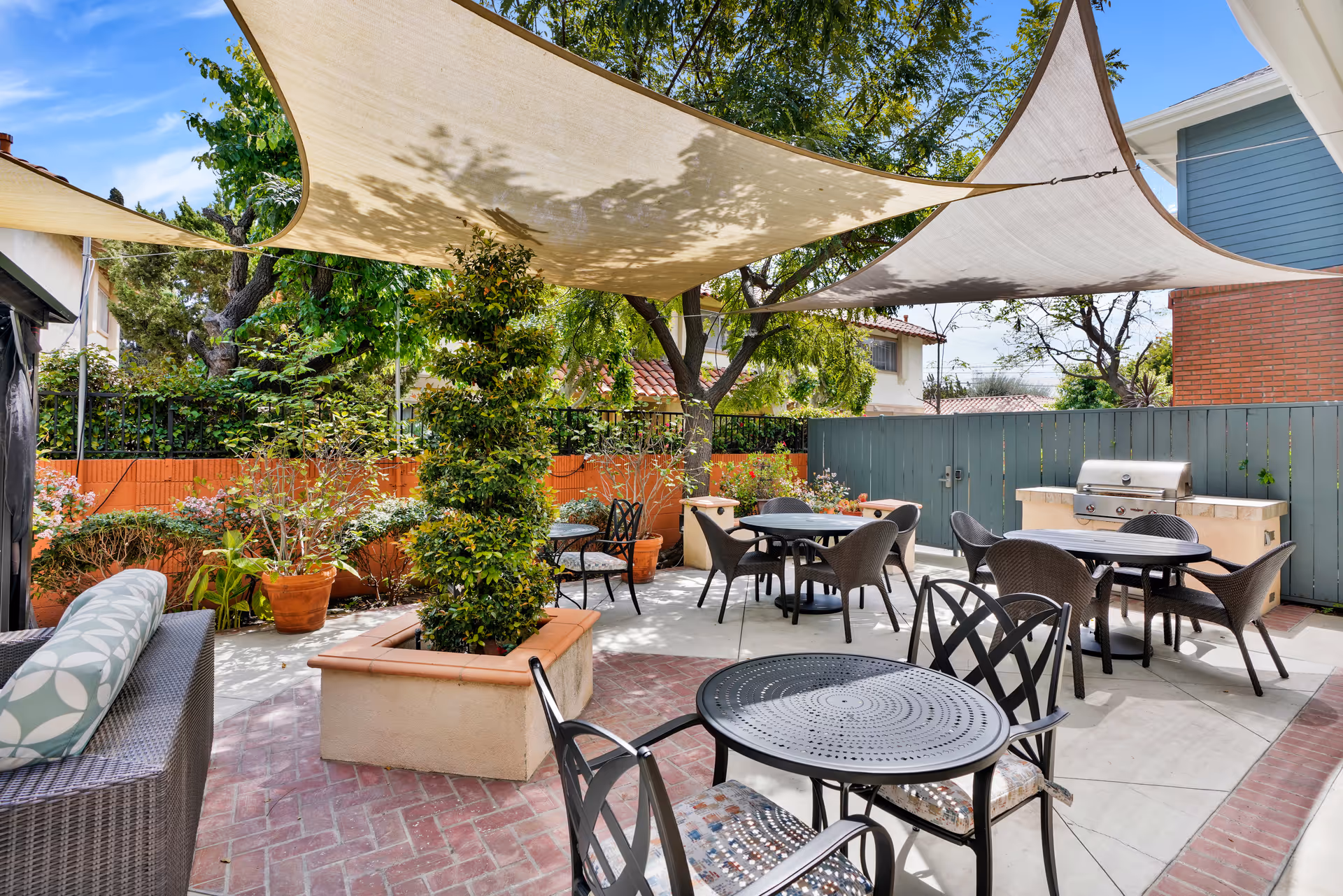 Outdoor patio area with several round tables and chairs, shaded by large beige fabric canopies. There are potted plants and greenery along the perimeter, a built-in barbecue grill, and a cushioned wicker bench on the left side.