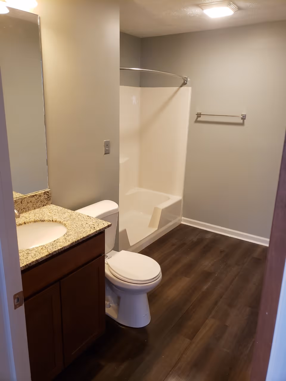 A clean bathroom featuring a white toilet, a bathtub with a built-in seat and curved shower rod, a granite countertop with an under-mount sink, wooden cabinetry, and a towel rack mounted on a light gray wall. The floor is covered with dark wood-style flooring.